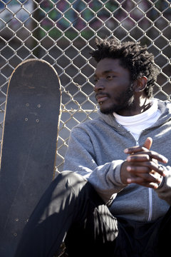 A serious young man leaning against a chain link fence next to a skateboard