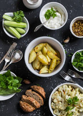 Baked cauliflower and potatoes, fresh cucumbers and herbs, sauces and spices on a dark stone background. Healthy vegetarian lunch or snack in a rustic style. Top view