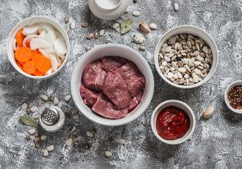 Raw ingredients - beef, beans, onions, carrots, spices and herbs, tomato sauce on a light stone background