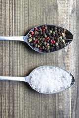 salt and pepper. Two metal spoons witlh salt crystals and color peppercorns on wooden table.