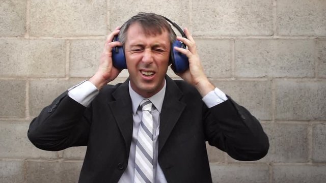 Man wearing a business suit and tie against a cinder-block wall with noise blocking safety acoustic earmuffs  grimacing in pain from loud noises.