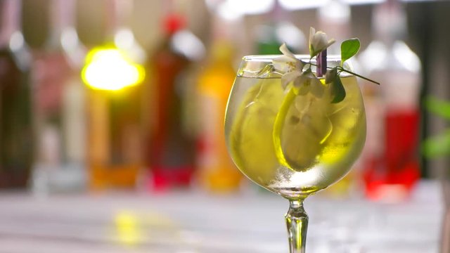 Glass of cocktail slowly rotates. Cocktail glass decorated with flower. Slices of lemon and ice. Chilled tom collins with lemon.