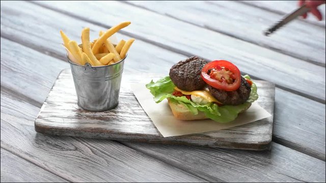 Hand with tongs prepares burger. French fries and hamburger. Grilled meat and fresh lettuce. Original fresh burger on table.