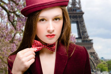 Portrait of elegant sexy girl in red velvet coat and hat holding bow tie near Eiffel tower