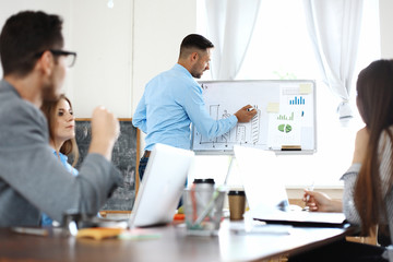 Handsome young man standing near whiteboard and pointing on the chart while his coworkers listening and sitting at the table