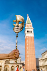 Fototapeta na wymiar Female hand holding Venetian carnival mask with San Marco tower on the background