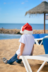 Back view of cute little boy in Santa hat on beach. Sunny exotic vacation outside