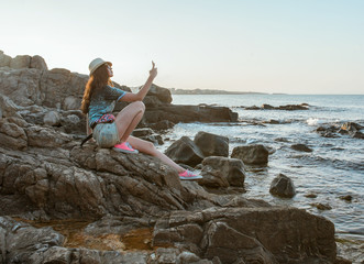 young attractive woman tourist making photo on mobile on rocky sea coast at sunset