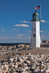 Fototapeta na wymiar American Flag Over Sunlit Historic Scituate Lighthouse Tower Along Rocky Shore in Massachusetts