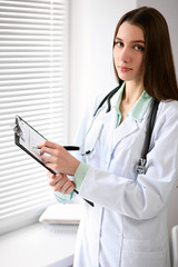 Female doctor brunette sitting  at the table near the window in hospital and typing at laptop computer
