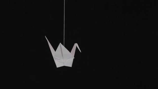 White paper origami crane swinging on thread against black background