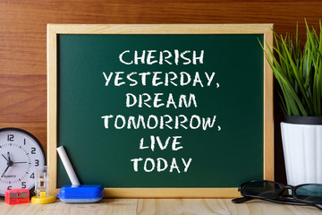 Word quote CHERISH YESTERDAY,DREAM TOMORROW,LIVE TODAY written on green chalk board on wooden table.