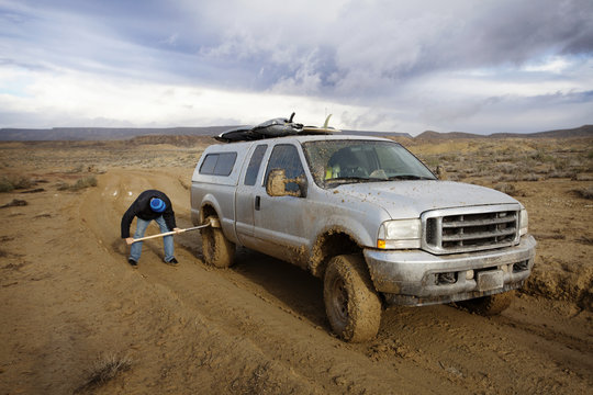 Man cleaning dirty tires of vehicle with shovel on land against cloudy sky