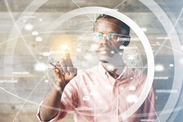 Portrait of African man in glasses, using digital device, pointing at icons on futuristic screen interface against high-tech interior, looking with serious concentrated expression. Selective focus