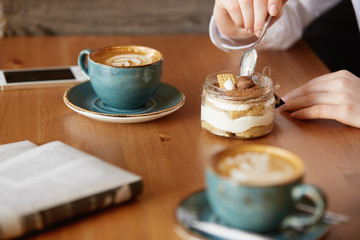 Close up shot of woman's hands eating sweet dessert with a spoon. Young female office worker having coffee with a colleague during lunch break while sitting at the wooden table at a restaurant. 