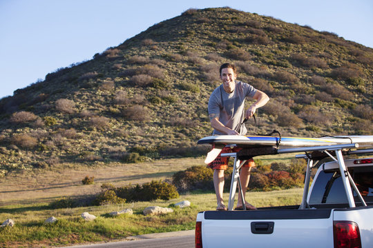 Smiling man tying surfboard on pick-up truck parked at roadside against hill