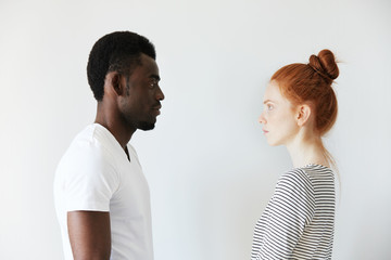 Fototapeta premium Sideways portrait of a couple looking each other in the eyes on white background. Afro man standing still and Caucasian redhead girl as well watching him with serious look like wife on husband.