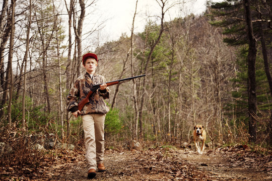 Boy holding rifle walking with dog on field in forest