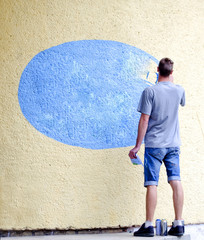 Young man paints blue oval on an external concrete wall. Adult male graffiti artist paints the wall.