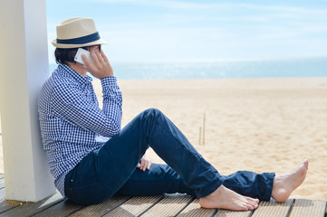 Fototapeta na wymiar Man in hat speaking on smartphone over blue sky and ocean beach background