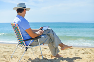 Man using tablet relaxing on ocean beach, blue sky outdoors background