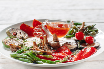 Grilled vegetables with red sauce on white plate. Front view on grilled asparagus, vegetable marrow, tomato, pepper, eggplant and mushrooms on white plate with cup of ketchup close-up