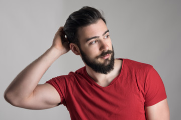 Young handsome bearded man in red t-shirt scratching head looking away over gray background