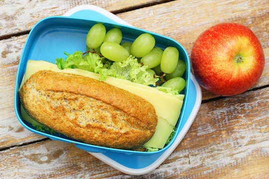 Healthy lunch box containing brown cheese roll, red apple and grapes on rustic wooden surface
