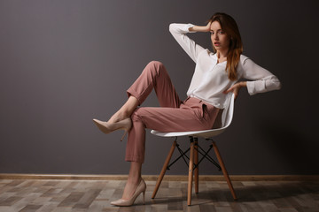 Beautiful young woman in shirt and pink pants sitting on chair on dark grey wall background