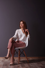 Beautiful young woman in shirt and pink pants sitting on chair on dark grey wall background