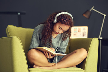 African American with headphones writing diary on green armchair