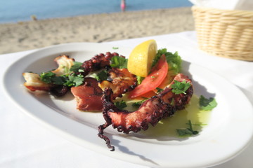 Grilled octopus with olive and a lemon slice on a plate by the sea. Grilled octopus legs on a tavern table by a Greek beach.