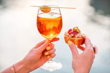 Female hand holding glass with Spritz Aperol alcohol drink decorated with olive on the water chanal...