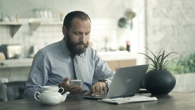 Young businessman comparing data on laptop and smartphone by table in kitchen
