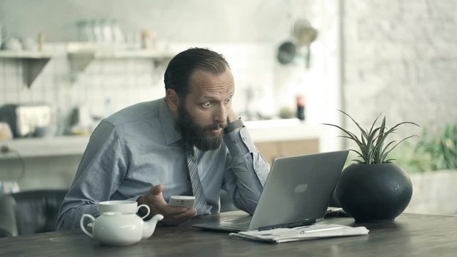 Sad, overwhelmed businessman working with laptop and smartphone in kitchen at home
