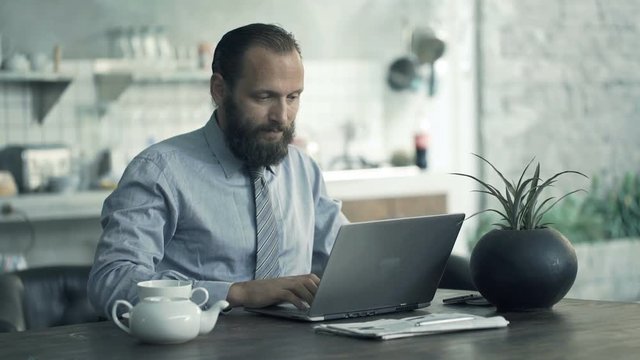 Young businessman working on laptop sitting by table in kitchen at home
