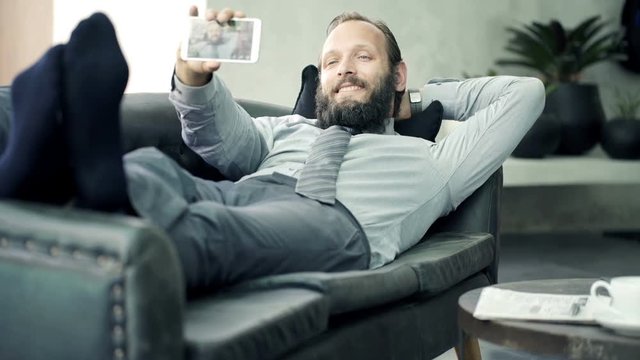 Young businessman taking selfie photo with cellphone lying on sofa at home
