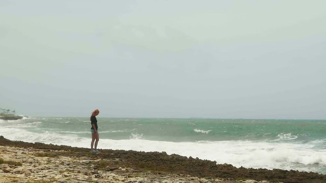 Attractive young girl with long red hair standing on the beach near the storm sea, slow-motion