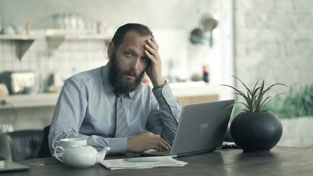 Portrait of sad, overwhelmed businessman with laptop and documents in kitchen
