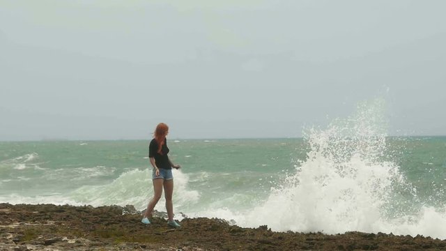 Attractive young woman with long red hair standing on the coast and looking to the storm sea, throws pebbles, slow-motion, wide angle