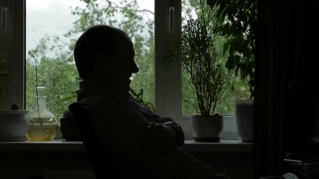 Silhouette of a young man in profile sitting against the window, and the window is bad weather and the wind is blowing. Static shooting.