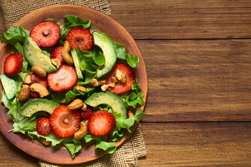 Strawberry, avocado, lettuce salad with cashew nuts on plate, photographed overhead on dark wood with natural light