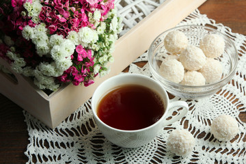 a Cup of tea and a glass bowl full of candies on a white napkin near the bouquet of beautiful and fragrant flowers in wooden box on wooden background