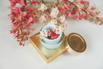 Fototapeta na wymiar There White and Pink Branches of Chestnut Tree,Goden Present Box with Bottle Cream are on White Table