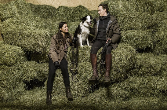 Full length rear view of happy young couple with dog sitting on hay stack in stable