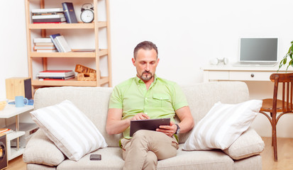 Handsome man sitting on sofa or couch and using tablet PC at home. Serious man working from home. Business or freelance concept.