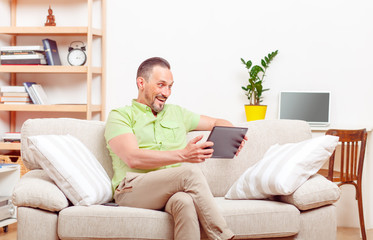 Portrait of handsome man sitting on sofa and using tablet PC at home for personal purposes: work, business, communication with foreign friends or business partners.