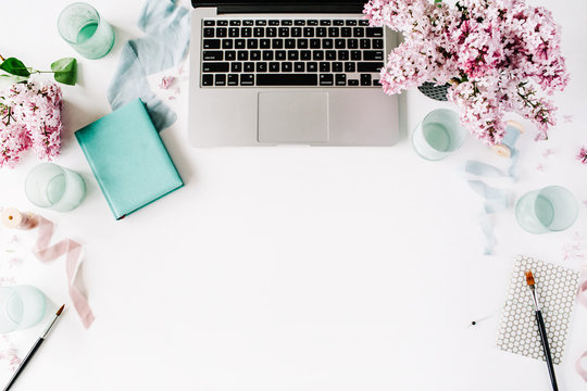 Workspace with paintbrush, laptop, lilac flowers bouquet, spool with beige and blue ribbon, mint diary on white background. Flat lay, top view