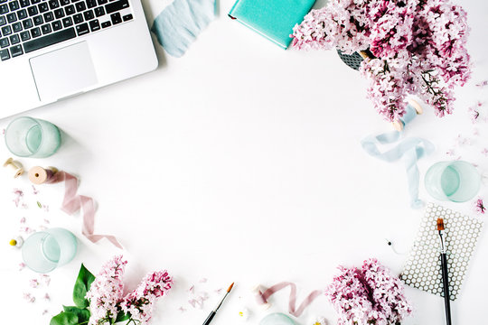 Workspace with paintbrush, laptop, lilac flowers bouquet, spool with beige and blue ribbon, mint diary on white background. Flat lay, top view