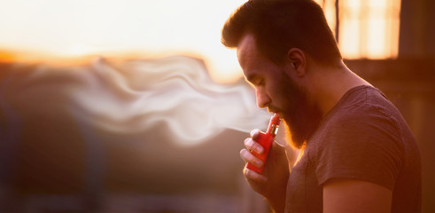 vaping, young man with a beard, produces vapor on sunset sky background, place for text